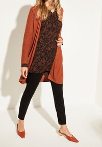 Langer kardinalroter Cardigan über einem schwarzen gemusterten Oberteil, kombiniert mit schwarzen Hosen und ruströtlichen Slippern. Glatte Textur und lässige Passform.