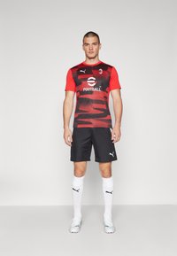 Camiseta de fútbol de color rojo y negro con estampado, pantalones cortos negros, calcetines blancos y botas con detalles en rosa. Logo de Puma en la camiseta y en los pantalones cortos.