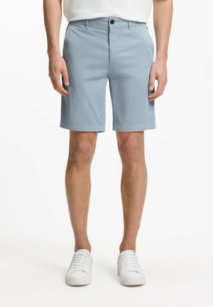 Man wearing light blue knee-length shorts with a button and zipper, white sneakers, and a white shirt, standing against a plain background.