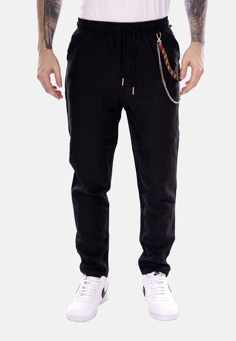 Uomo che indossa pantaloni jogger neri con un accessorio a catena intrecciata, sneaker bianche e una maglietta bianca, in piedi davanti a uno sfondo semplice.
