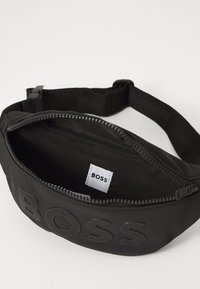 Sac banane noir avec une finition texturée, doté d'une fermeture éclair et d'une étiquette intérieure avec le logo "BOSS". Sangle ajustable pour plus de confort.