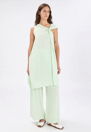 Donna con lunghi capelli castani in piedi che indossa una tunica verde chiaro senza maniche con dettaglio nodo, pantaloni larghi coordinati e scarpe basse bianche.