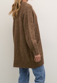Mujer con un largo cárdigan de punto marrón con puños y dobladillo acanalados, combinado con jeans azules, de pie de lado contra un fondo sencillo.