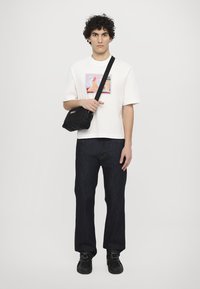 Witte t-shirt met kleurrijk grafisch ontwerp, donkerblauwe spijkerbroek en zwarte crossbody tas. Model staat met de handen op de tas geplaatst.
