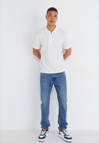 Camisa polo blanca con cuello, combinada con jeans de mezclilla azules. Zapatillas blancas con acentos en azul y rojo. Estilo minimalista.