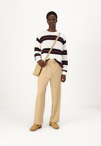 Suéter blanco con rayas negras y rojas, pantalones color beige y zapatillas color miel. Al lado del modelo descansa un bolso cruzado beige.