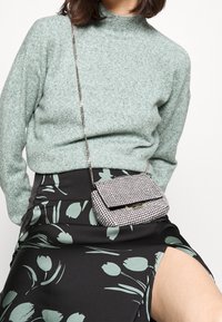 Žena ve světle zeleném svetru, černé sukni s zeleným květinovým vzorem, s vysokým rozparkem a malou stříbrnou kabelkou na řetízku přes tělo.