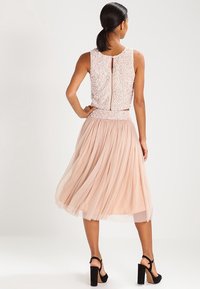 Kleid mit einem ärmellosen, mit Pailletten besetzten Oberteil und einem Reißverschluss, kombiniert mit einem fließenden, plissierten, hellrosa Rock und schwarzen High-Heels.