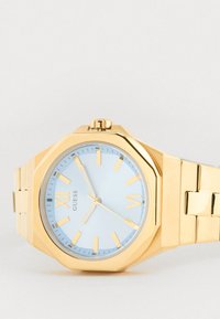 Orologio da polso in metallo dorato con quadrante ottagonale, fondale blu chiaro, indicatori delle ore dorati e bracciale a maglie dorate. Presenta una corona per la regolazione.