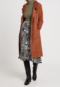 Brauner Trenchcoat aus Wildleder über einem grünen Rollkragenpullover, kombiniert mit einem gestuften Rock mit Zebramuster und kniehohen schwarzen Stiefeln.