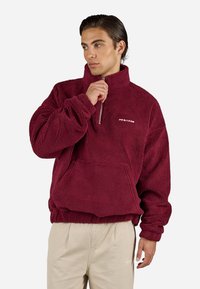 Mladic u bež hlačama nosi burgundski flis pulover s polu-zip ovratnikom i prednjim kengurudžijom džepom, gleda u stranu.