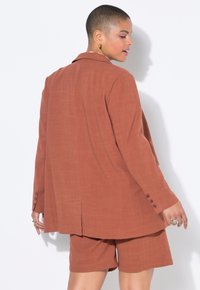 Lněné sako rezavé barvy s uvolněným střihem, jedním knoflíkem a odpovídajícími šortkami, vyznačující se hladkou texturou a minimalistickým designem.