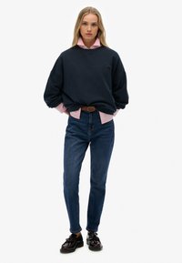 Un sweat-shirt navy superposé à une chemise à col rose clair, associé à un jean bleu foncé et des chaussures noires avec un détail en gland.