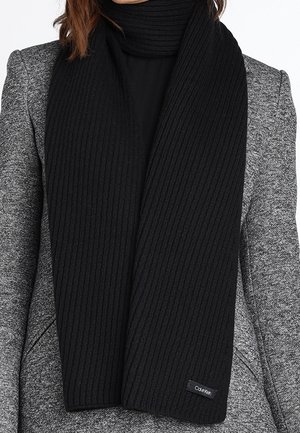 Femme portant une écharpe en maille côtelée noire sur un haut noir et un blazer gris texturé, écharpe étiquetée Calvin Klein.