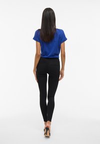 Top corto de manga corta azul para mujer combinado con jeans ajustados negros y zapatos de tacón altos negros, visto desde atrás sobre un fondo blanco.