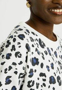 Primer plano de una persona sonriente que lleva un suéter blanco con estampado de leopardo negro y azul y un pendiente dorado en forma de aro retorcido.