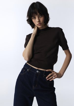 Giovane con capelli corti e scuri che posa indossando una maglietta nera a maniche corte e jeans blu scuro a vita alta su uno sfondo semplice.
