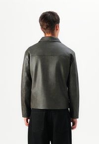 Dark green leather jacket with a structured collar and long sleeves. Smooth texture, fitted cut, and minimalist design, worn over black trousers.