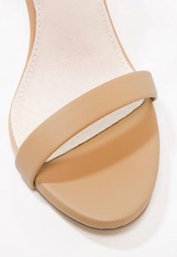 Beige Sandale mit offenen Zehen, die einen breiten Riemen vorne hat und eine weiche, gepolsterte Innensohle mit sichtbarer Naht.