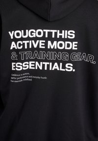 Melns kapučjakas ar lielu baltu tekstu: "TEV TAS IZDOSIES AKTĪVAIS REŽĪMS UN TRENIŅA APRĪKOJUMS. PAMATLĪDZĪBAS." Gluda auduma tekstūra ar vaļīgu piegriezumu.