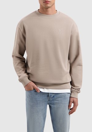 Beige sweatshirt with round neckline, ribbed cuffs and hem, featuring a small embossed logo on the chest, paired with light blue jeans.
