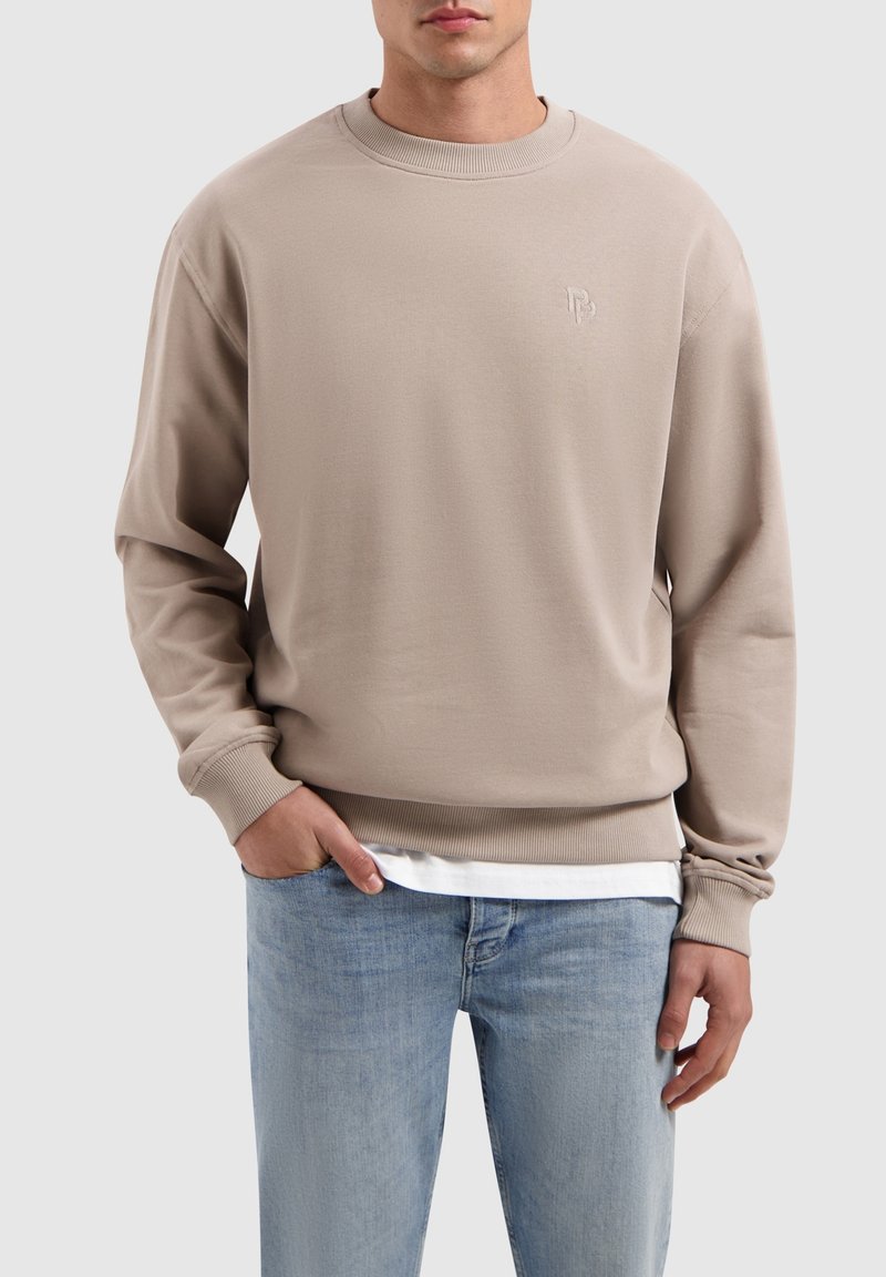 Beige sweatshirt met ronde hals, ribbenmanchetten en -zoom, met een klein reliëf logo op de borst, gecombineerd met lichtblauwe jeans.