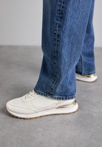 Sneaker bianco con tomaia in pelle texturizzata, accentuazione dorata sul tallone e suola in gomma bianca, abbinato a jeans blu sbiaditi in un contesto casual.