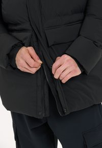 Musta puhvijakku, jossa on vetoketju ja napit, sileällä pinnalla ja vetoketjullinen sivutasku lämpötilan säilyttämiseksi.