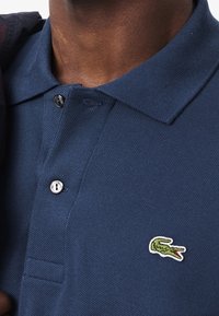 Polo-shirt marine en tissu texturé, avec deux boutons et un petit logo de crocodile vert sur la poitrine gauche.