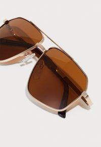 Gafas de sol de tono marrón con un marco de metal dorado, lentes cuadrados y acentos texturizados en las patillas, con almohadillas nasales transparentes.