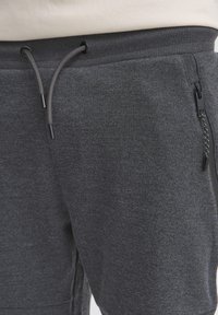 Pantalone za treniranje u boji ugljena s rebrastim pojasom, zatvaranjem na vezicu i džepovima sa zatvaračem, izrađeni od mekog, teksturiranog materijala.