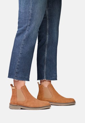 Personne portant un jean bleu court et des bottines Chelsea en daim marron clair, debout sur un fond clair uni.