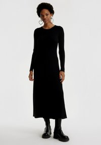 Schwarzes, geripptes, langärmeliges Kleid mit Rundhalsausschnitt, präsentiert in einer geraden Silhouette und Seitenschlitzen, kombiniert mit schwarzen Plateauboots.