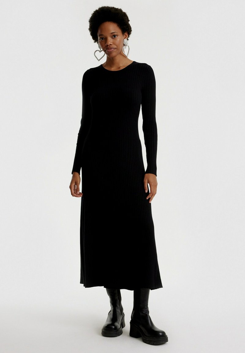 Schwarzes, geripptes, langärmeliges Kleid mit Rundhalsausschnitt, präsentiert in einer geraden Silhouette und Seitenschlitzen, kombiniert mit schwarzen Plateauboots.