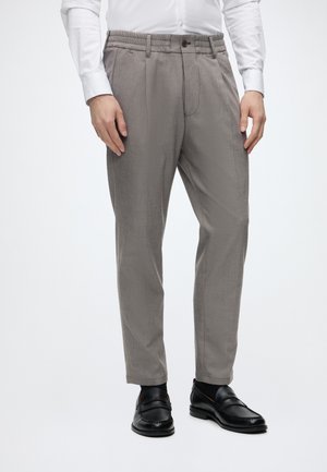 Homme portant un pantalon gris clair sur mesure, des mocassins en cuir noirs et une chemise blanche, debout devant un fond uni.