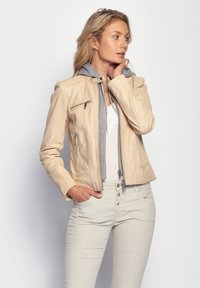 Lädernjacka i ljusbeige med en grå hoodie underneath. Har dragkedjestängning, kviltade axeldetaljer och framfickor.