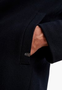 Marineblå tekstureret jakke med en synlig hånd i lommen. Har en metallisk logomærke nær lommens søm. Nærbillede af stof og detalje.