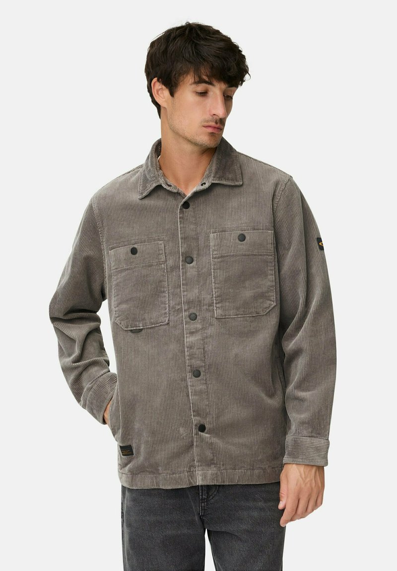 Man in een grijze corduroy knopenjack met twee borstzakken en zwarte jeans, poserend met één hand in de zak.
