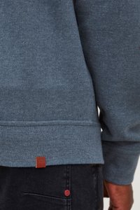 Blå hettegenser med ribbestrikkede mansjetter og nederkant, med et lite brunt lærlogo-merke. Glatt tekstur, uformell design. Mørke jeans er delvis synlige.