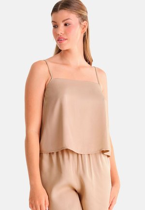 Vrouw draagt een beige mouwloze losse top met dunne bandjes en bijpassende beige broek, kijkt iets naar rechts.