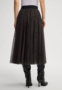 Mulher a usar uma saia midi com padrão preto e castanho, com um cós elástico preto e botas de couro de salto alto pretas, em pé.