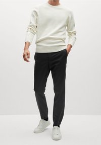 Jasno beżowy sweter dzianinowy z prążkowanymi mankietami, zestawiony z czarnymi spodniami i białymi trampkami. Sweter ma klasyczny okrągły dekolt.