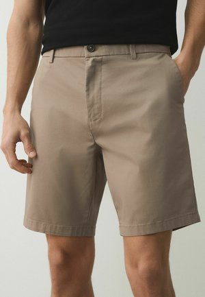 Bărbat purtând pantaloni scurți de culoare bej până la genunchi cu buzunare laterale și o cămașă neagră băgată în pantaloni, stând cu mâinile relaxate.