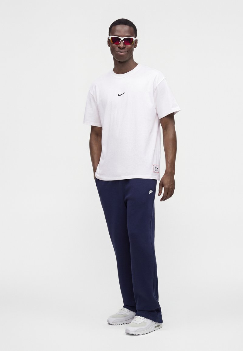 Mand iført rødtonede sportssolbriller, hvid Nike t-shirt, mørkeblå Nike bukser og hvide sneakers, står mod en ensfarvet baggrund.