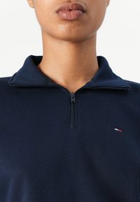 Marineblå zip-up krageplagg laget av myk stoff, med en liten brodert logo i rødt, hvitt og blått på brystet.