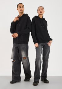 To modeller har på seg svarte hettegensere og slitte svarte jeans. Hettegenserne har snorer, mens jeansene har strategiske hull på lårene.