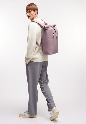 Persona giovane con maglione color crema e pantaloni grigi che porta uno zaino alto color malva con tasca frontale, in piedi su sfondo bianco.