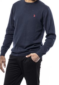 Mörkblåstickad tröja med långa ärmar och rund halsringning. Har en liten röd logotyp på vänster bröst. Matchas med svarta byxor.