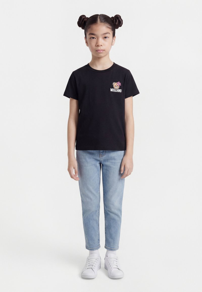 Νεαρή κοπέλα στέκεται προς τα εμπρός φορώντας μαύρο t-shirt Moschino με λογότυπο αρκουδάκι, ανοιχτό μπλε τζιν, λευκές κάλτσες και λευκά παπούτσια.