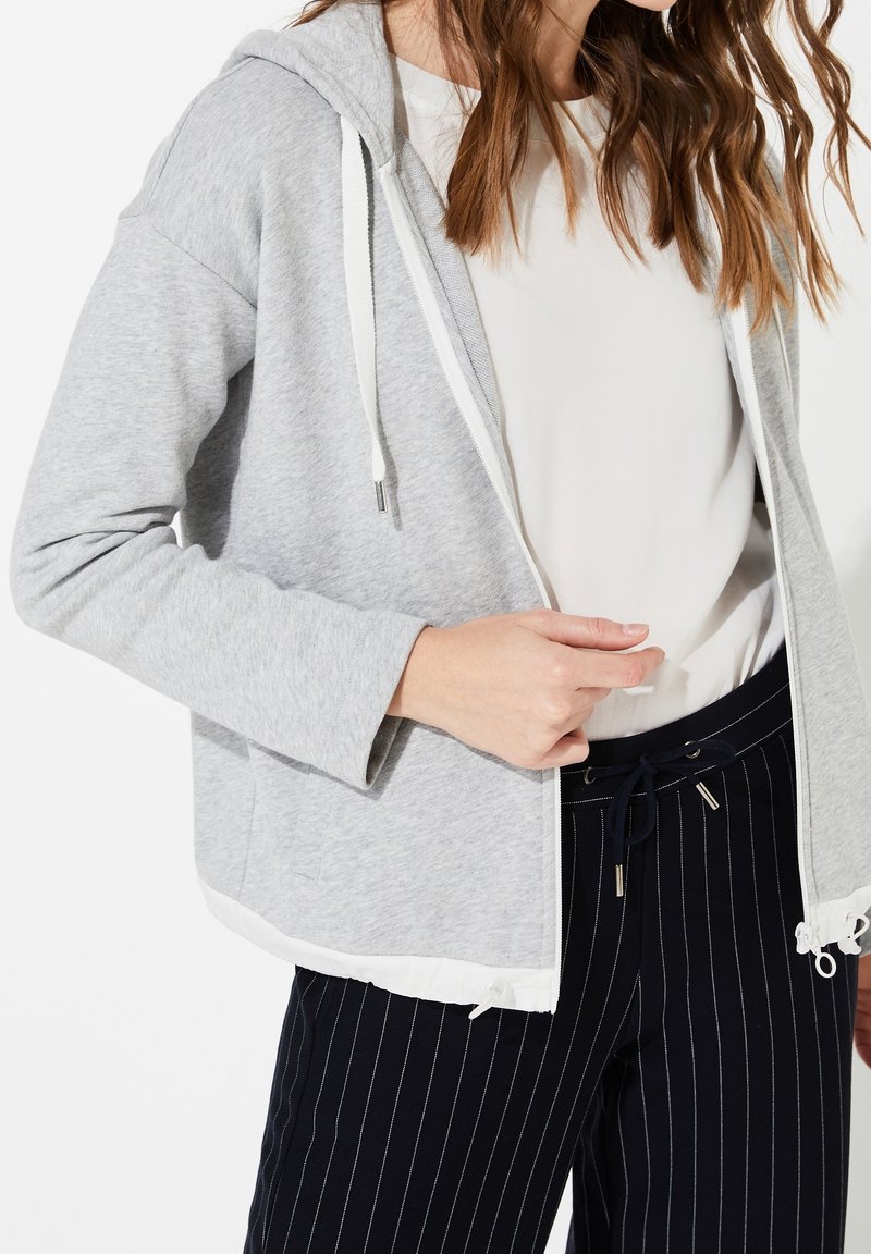 Sudadera gris con capucha y cordones blancos, con un dobladillo blanco y un corte relajado, combinada con pantalones azul marino a rayas finas.